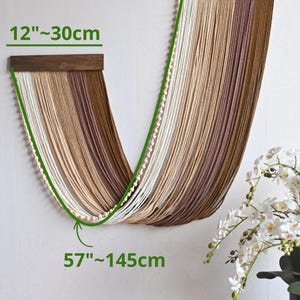 May include: A wall hanging featuring two dark wooden bars with draped, multi-toned brown and cream yarn strands. The piece includes a string of small wooden beads along the edge. Dimensions are 30 cm wide and 145 cm long.
