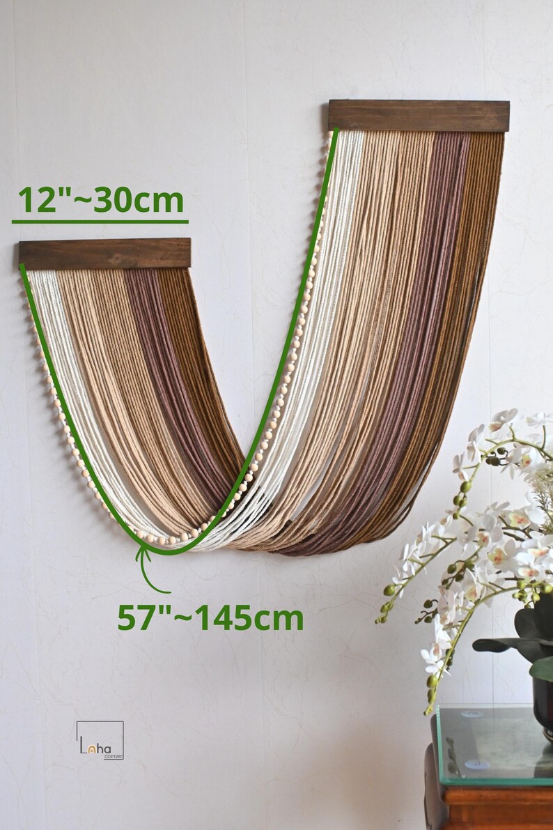 May include: A wall hanging featuring two dark wooden bars with draped, multi-toned brown and cream yarn strands. The piece includes a string of small wooden beads along the edge. Dimensions are 30 cm wide and 145 cm long.