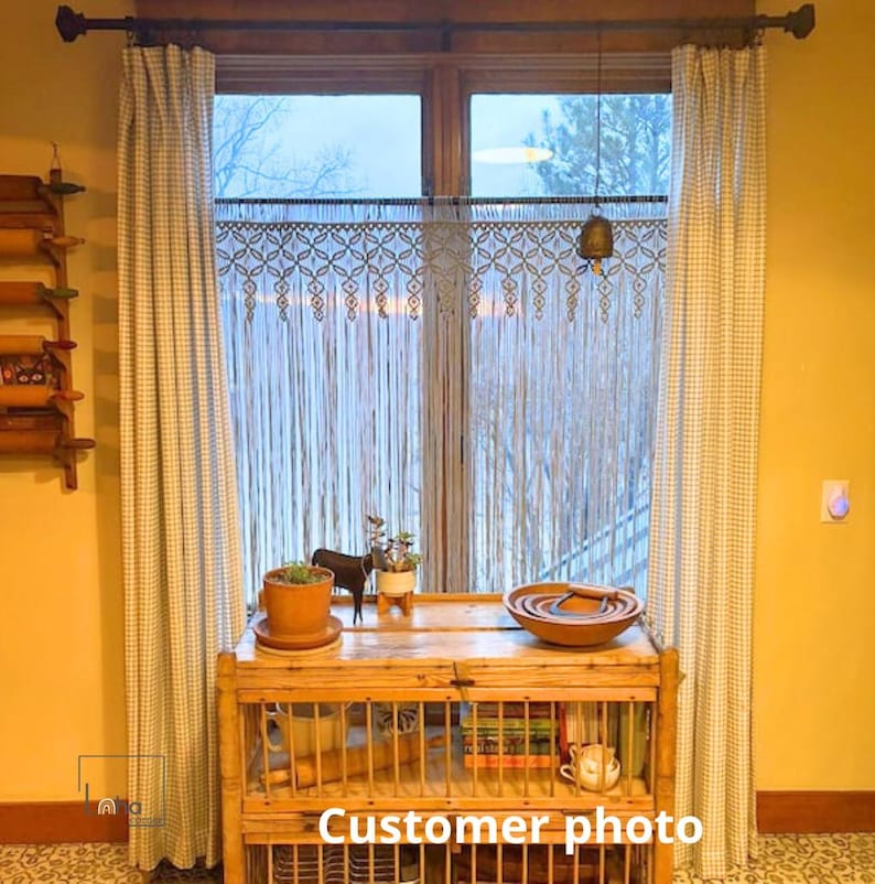 May include: A window featuring light-colored curtains with a small grid pattern, a macrame valance with a geometric fringe, and a wooden console table holding a terracotta pot, a small animal figurine, and a wooden bowl with nested items.
