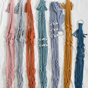May include: Six macrame plant hangers in different colors, including pink, gray, orange, teal, light blue, and dark blue. Each hanger has a wooden bead accent and a metal ring at the top.