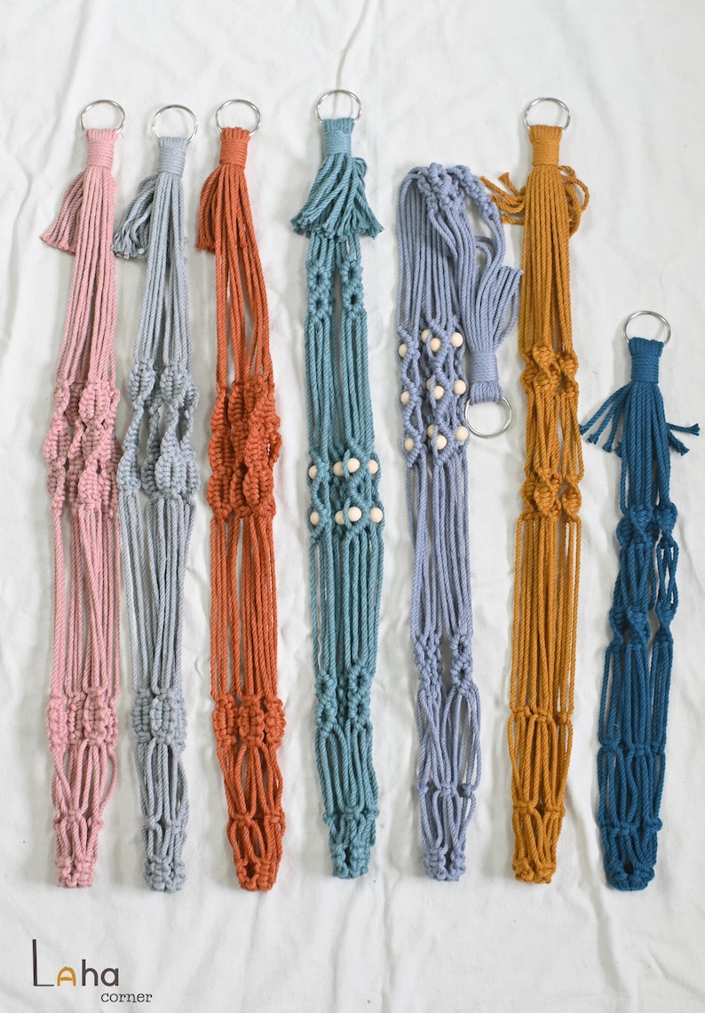 May include: Six macrame plant hangers in different colors, including pink, gray, orange, teal, light blue, and dark blue. Each hanger has a wooden bead accent and a metal ring at the top.