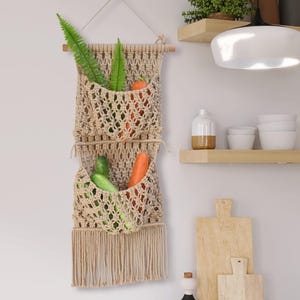 以下が含まれることがあります： ベージュのマクラメの壁掛け。野菜と緑が入った2つのポケットがあります。織りのデザインは下部にフリンジがあり、木のダボと天然のロープで吊り下げられています。