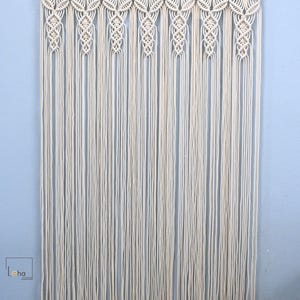 May include: A cream white macrame wall hanging suspended from a wooden rod, featuring intricate knotted patterns at the top and long, vertical fringe strands. The dimensions are 28 inches wide by 53 inches long, with the text Loha Corner visible.