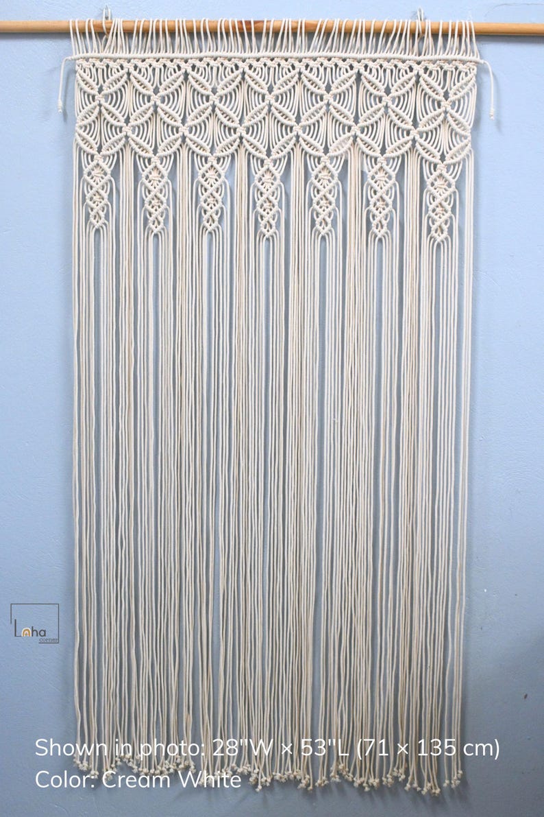 May include: A cream white macrame wall hanging suspended from a wooden rod, featuring intricate knotted patterns at the top and long, vertical fringe strands. The dimensions are 28 inches wide by 53 inches long, with the text Loha Corner visible.