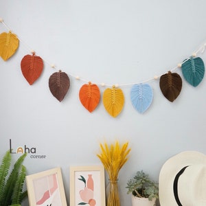 Peut inclure: Une guirlande de feuilles en macramé colorée avec des feuilles brunes, orange, jaunes, bleues et vertes. La guirlande est enfilée sur une ficelle blanche et des perles en bois. La guirlande est suspendue à un mur bleu clair.