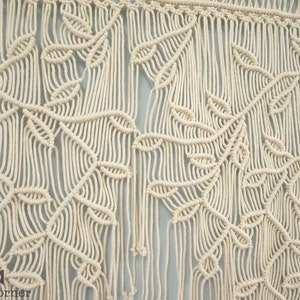 May include: A cream-colored macrame wall hanging featuring intricate knotted patterns that form repeating leaf and vine motifs. The design includes long, vertical fringe strands hanging from a horizontal top bar, with the text Laha corner visible.