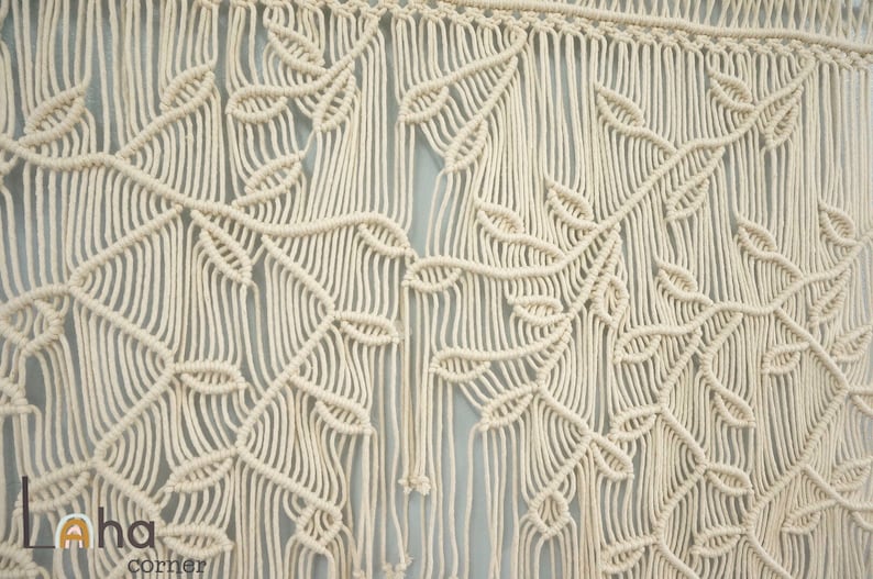 May include: A cream-colored macrame wall hanging featuring intricate knotted patterns that form repeating leaf and vine motifs. The design includes long, vertical fringe strands hanging from a horizontal top bar, with the text Laha corner visible.