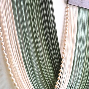 May include: A wall hanging featuring long, vertical strands of yarn in shades of cream, sage green, and dark forest green. The yarn is suspended from a dark brown wooden bar and accented with two vertical strings of small, round, light-coloured wooden beads.