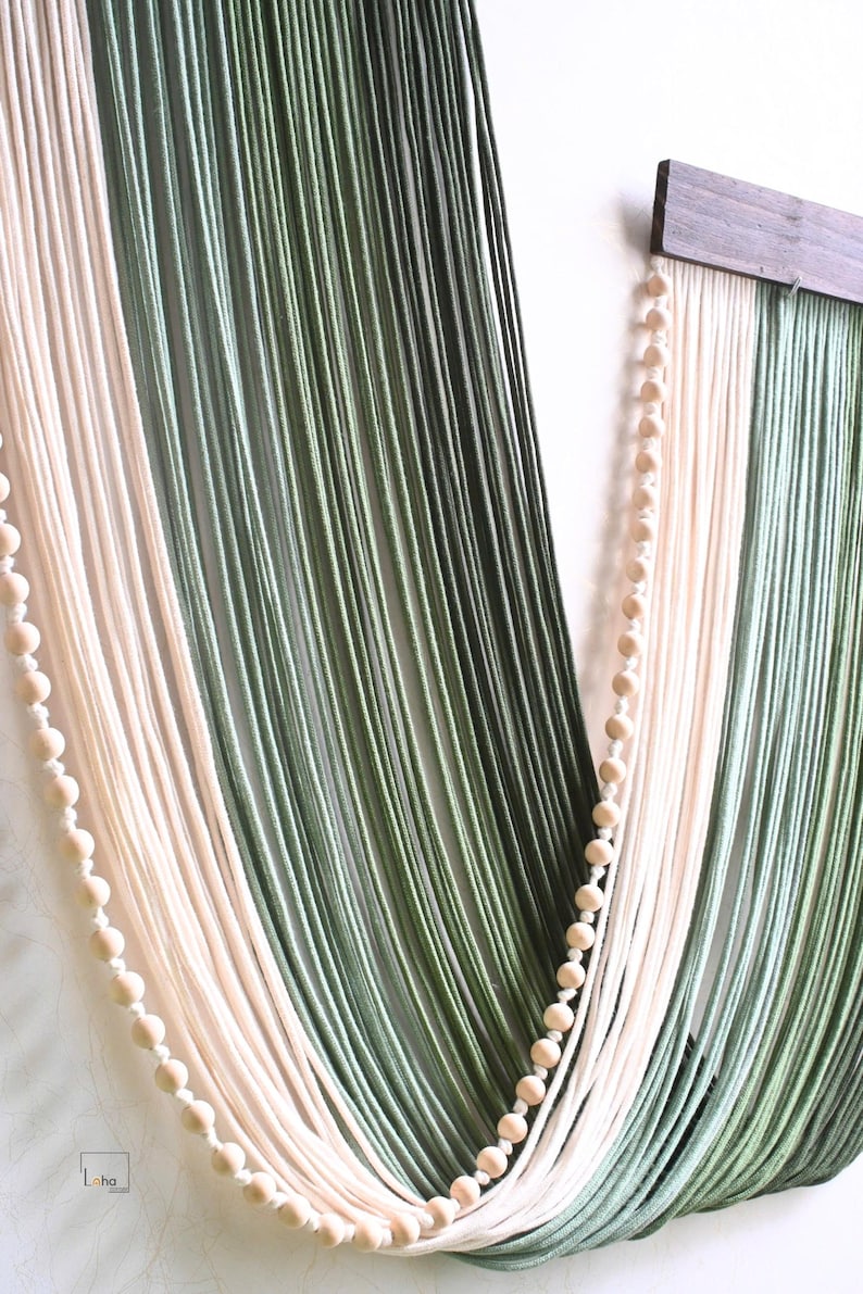 May include: A wall hanging featuring long, vertical strands of yarn in shades of cream, sage green, and dark forest green. The yarn is suspended from a dark brown wooden bar and accented with two vertical strings of small, round, light-coloured wooden beads.