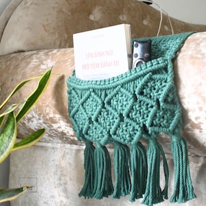May include: A sage green macrame hanging organiser featuring a diamond knot pattern and five decorative tassels at the bottom. The organiser holds a white book with the text LÊN ĐỈNH NÚI MỞ TIỆM BÁNH MÌ and a black remote control in its pocket.