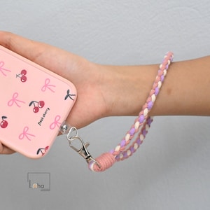 Puede incluir: Funda de teléfono rosa con un estampado de cerezas y lazos, sujeta a una correa de muñeca trenzada. La correa es una mezcla de hilos rosas, morados y blancos, asegurada con un cierre plateado.