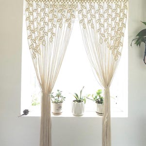 May include: A pair of white macrame curtains with a leaf pattern hanging in a window. The curtains are made of thick cotton cord and are tied back with a simple knot. There are three potted plants on the windowsill.
