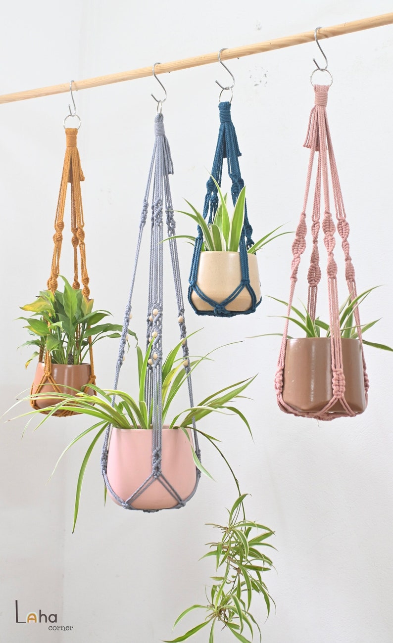 May include: Four macrame plant hangers with different colored cords and wooden beads. The hangers are holding potted plants. The hangers are hanging from a wooden dowel rod.
