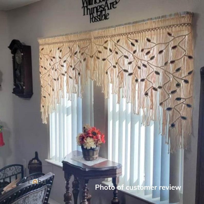 May include: A cream-colored macrame window valance featuring an intricate knotted branch pattern with dark leaf-shaped accents. The valance has a long, layered fringe edge and hangs across the top of a window with vertical blinds.