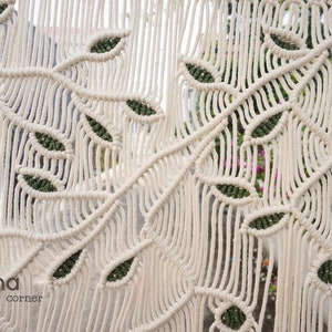 May include: A macrame wall hanging featuring off-white cotton cord knotted into a repeating branch and leaf pattern with dark green accents within the leaves. The text Laha corner is printed in the lower left corner of the textile piece.