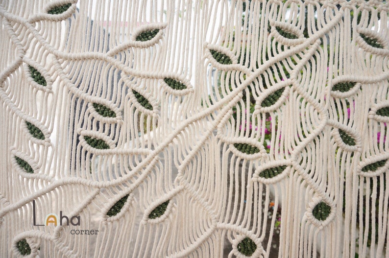 May include: A macrame wall hanging featuring off-white cotton cord knotted into a repeating branch and leaf pattern with dark green accents within the leaves. The text Laha corner is printed in the lower left corner of the textile piece.