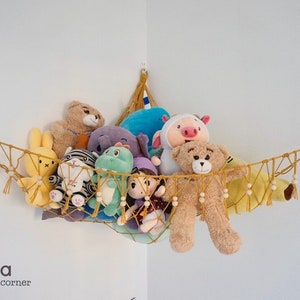 May include: A yellow macrame hammock with wooden beads hangs from a corner of a white wall. The hammock is filled with stuffed animals, including a teddy bear, a pig, a rabbit, a zebra, a dinosaur, and a doll. The hammock is decorated with rainbow-colored yarn.