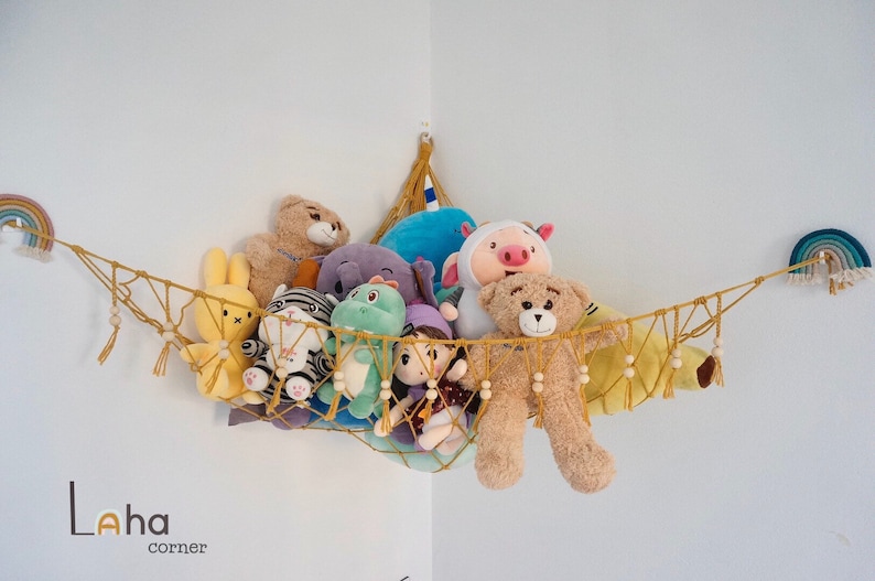 May include: A yellow macrame hammock with wooden beads hangs from a corner of a white wall. The hammock is filled with stuffed animals, including a teddy bear, a pig, a rabbit, a zebra, a dinosaur, and a doll. The hammock is decorated with rainbow-colored yarn.