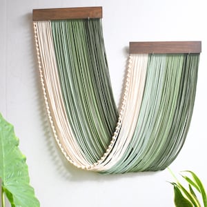 May include: A wall hanging featuring two dark wood horizontal bars from which long, draped strands of cream and sage green cord are suspended in a U-shape. A single row of small, round, light-coloured wooden beads accents the cream-coloured cord section.