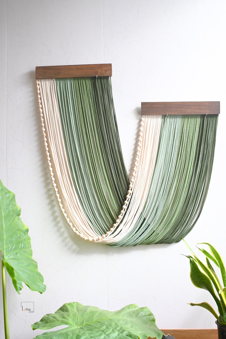 May include: A wall hanging featuring two dark wood horizontal bars from which long, draped strands of cream and sage green cord are suspended in a U-shape. A single row of small, round, light-coloured wooden beads accents the cream-coloured cord section.