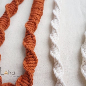Puede incluir: Dos juegos de cordones de macram&eacute; trenzados, uno en un color naranja quemado intenso y otro en un color crema blanquecino. Los cordones presentan un patr&oacute;n repetitivo de nudos en espiral a lo largo de su longitud. El texto Laha corner est&aacute; impreso.