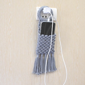 May include: A grey macrame phone holder featuring a circular top loop that hangs over a wall power adapter. The holder has a diamond-patterned woven body, a black smartphone inside, and four decorative tassels at the bottom. Text reads Laha corner.