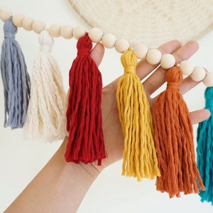 May include: A colorful tassel garland with wooden beads. The tassels are in shades of gray, white, red, yellow, orange, and teal.