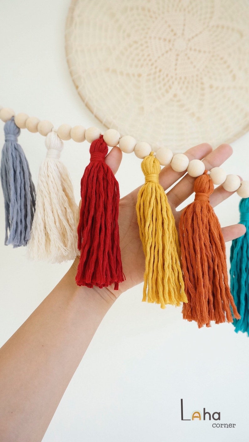 May include: A colorful tassel garland with wooden beads. The tassels are in shades of gray, white, red, yellow, orange, and teal.