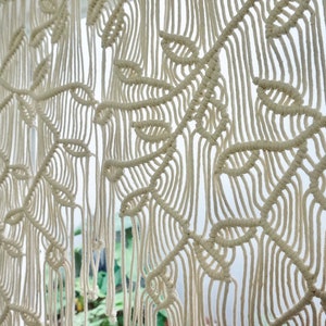 May include: An off-white macrame wall hanging featuring a repeating leaf pattern created with intricate knotted cord work. The design consists of vertical strands and woven foliage shapes that form a textured, hanging textile piece.