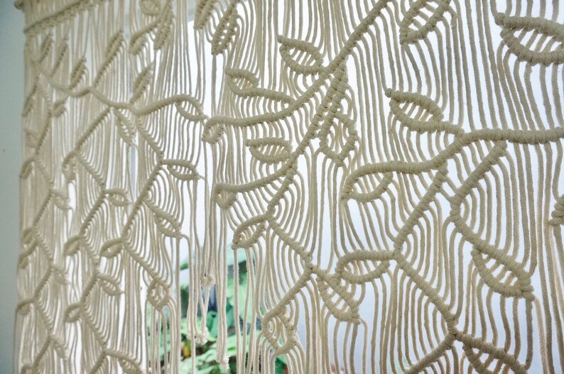 May include: An off-white macrame wall hanging featuring a repeating leaf pattern created with intricate knotted cord work. The design consists of vertical strands and woven foliage shapes that form a textured, hanging textile piece.