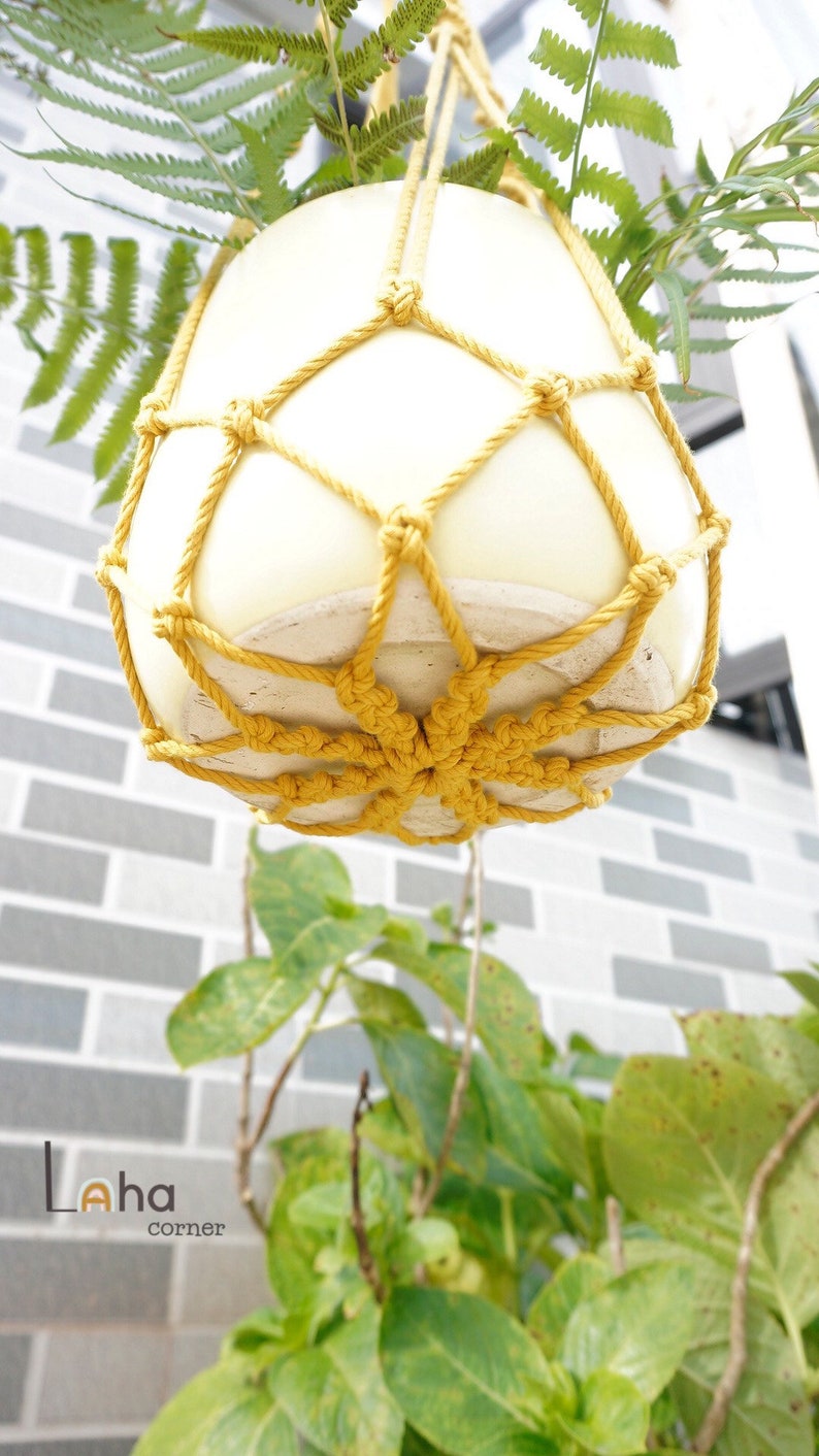 Pu&ograve; includere: Un vaso di ceramica bianco con un portavasi in macram&egrave; giallo. Il vaso &egrave; appeso al soffitto e ci sono foglie verdi nel vaso.