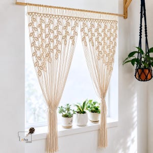 May include: A cream-colored macrame curtain featuring a repeating leaf-like knot pattern, suspended from a wooden rod. The curtain is divided into two panels, each gathered with a tie, and displays the Laha Corner logo in the bottom left corner.