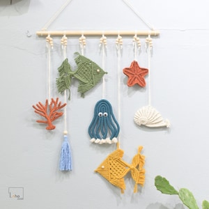 May include: A wall hanging with a nautical theme featuring a variety of crocheted sea creatures, including a green fish, a blue octopus, a yellow fish, a white whale, a red starfish, and a coral branch. The hanging is made with white string and has wooden beads.