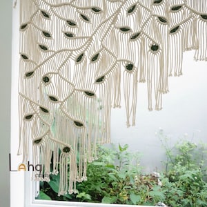 May include: A cream-colored macrame curtain hanging from a wooden rod, featuring an intricate knotted pattern that forms leaf-like shapes with dark green accents. The design includes long, dangling fringe at the bottom and the text Lahai home.