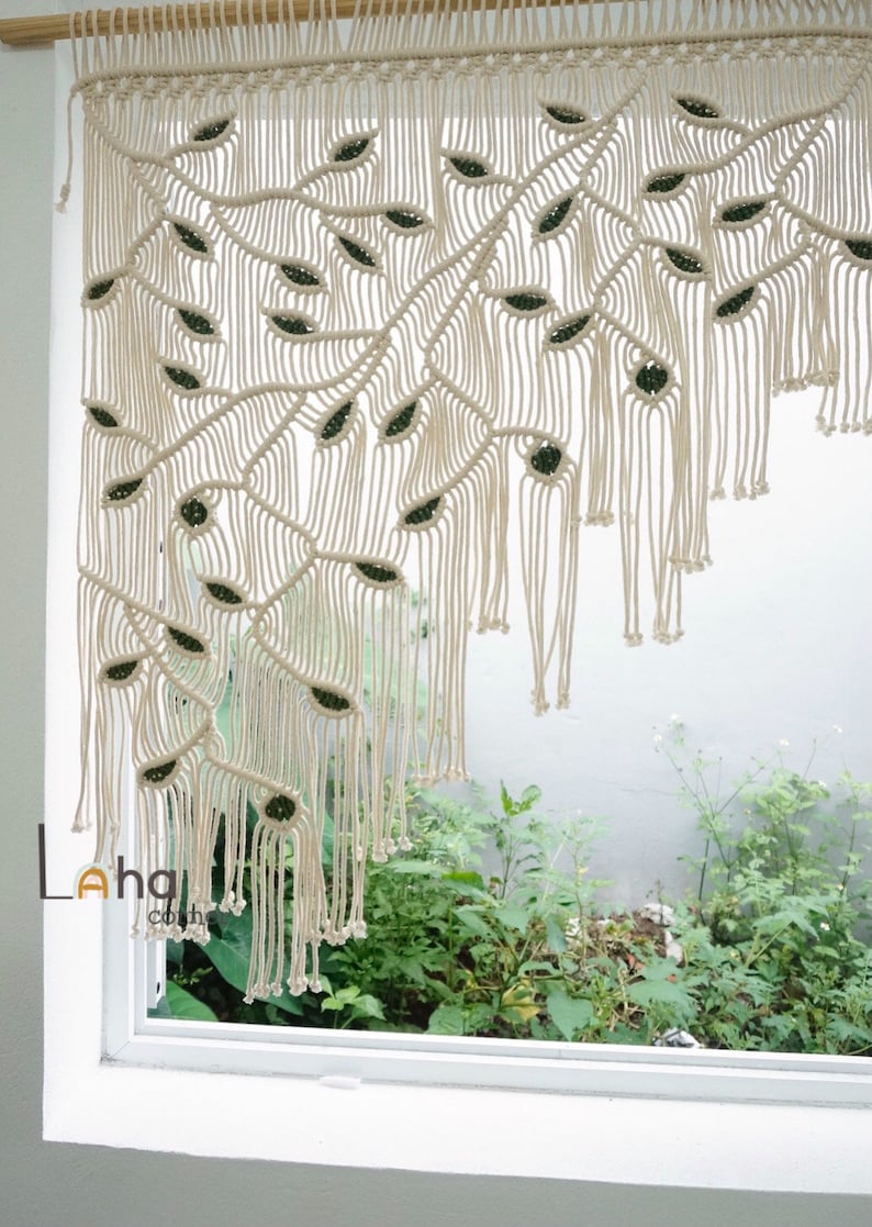May include: A cream-colored macrame curtain hanging from a wooden rod, featuring an intricate knotted pattern that forms leaf-like shapes with dark green accents. The design includes long, dangling fringe at the bottom and the text Lahai home.