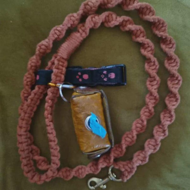 Puede incluir: Una correa para perro de macram&eacute; en color rosa empolvado con nudos en espiral, un collar negro con motivos de huellas rosas y un dispensador de bolsas de cuero color mostaza con un rollo de bolsas azul brillante, con herrajes met&aacute;licos.