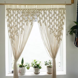 May include: A cream-colored macrame curtain featuring a repeating leaf-like knot pattern suspended from a wooden rod. The curtain is gathered at the sides, revealing a window with three potted plants on the sill and a hanging plant nearby.