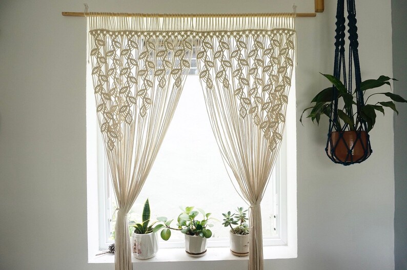 May include: A cream-colored macrame curtain featuring a repeating leaf-like knot pattern suspended from a wooden rod. The curtain is gathered at the sides, revealing a window with three potted plants on the sill and a hanging plant nearby.