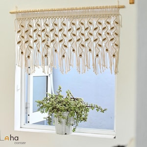 May include: A cream-coloured macrame window valance featuring a repeating leaf-like knot pattern with integrated brown wooden beads, suspended from a light-toned wooden rod. The intricate fibre art design hangs above a window with a green plant.
