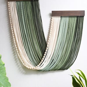 May include: A wall hanging featuring two dark brown wooden bars from which long, draped strands of cream and sage green cord hang in a U-shape. A single string of small, round, light-coloured wooden beads follows the curve of the draped cord.