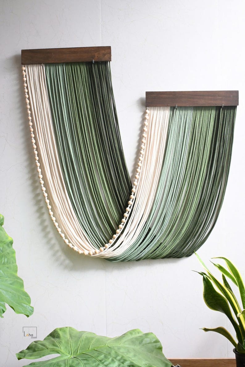May include: A wall hanging featuring two dark brown wooden bars from which long, draped strands of cream and sage green cord hang in a U-shape. A single string of small, round, light-coloured wooden beads follows the curve of the draped cord.