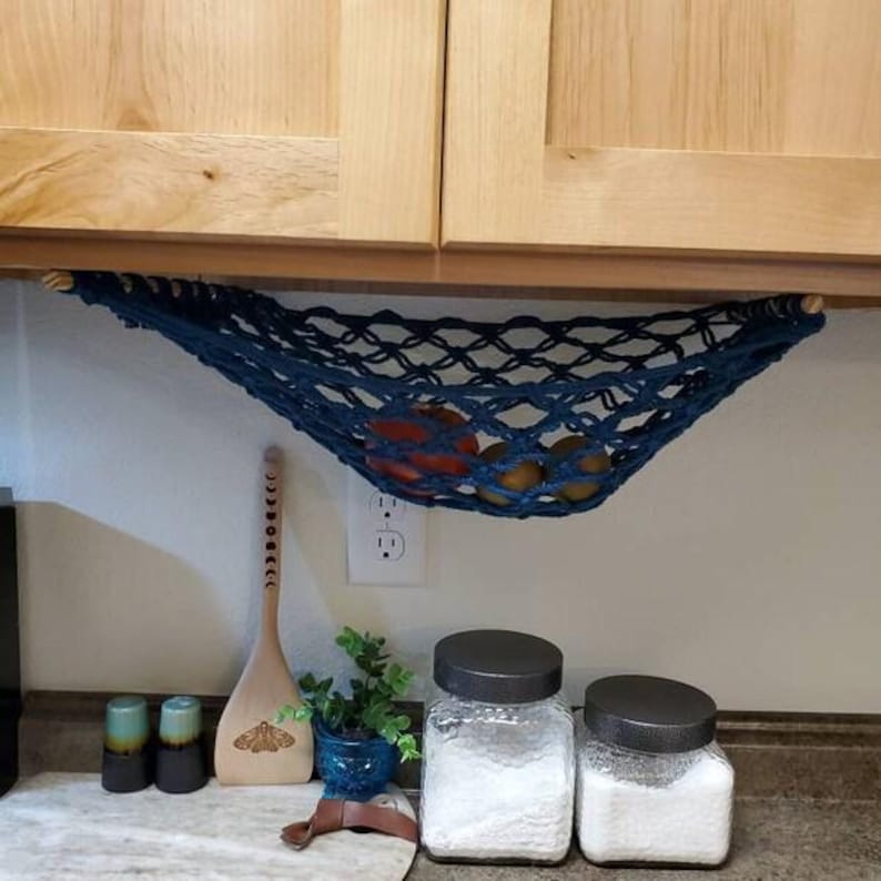 Puede incluir: Una hamaca de frutas de macram&eacute; azul marino suspendida debajo de armarios de madera, que contiene naranjas y otras frutas. La decoraci&oacute;n de la cocina incluye frascos, una cuchara de madera y peque&ntilde;as plantas en macetas. La hamaca mide unos 61 cm de ancho.