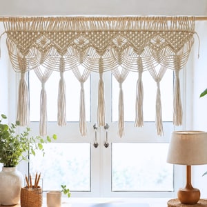 Peut inclure: Un rideau court en macramé de couleur crème présentant un motif complexe de losanges noués sur le haut, se prolongeant par des cordons drapés en cascade qui se terminent par huit longs pompons épais suspendus à une tringle en bois.
