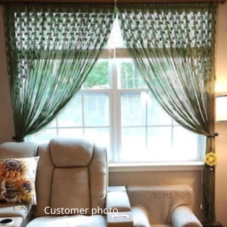 May include: A pair of olive green string curtains featuring a woven lattice pattern at the top header. The curtains hang vertically in front of a window, with the fabric strands draped to the sides and secured with circular yellow tiebacks.