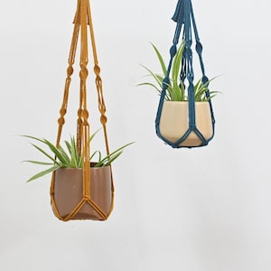 May include: Two macrame plant hangers, one in mustard yellow and one in teal blue, hanging from a wooden dowel rod. Each hanger has a small potted plant inside.