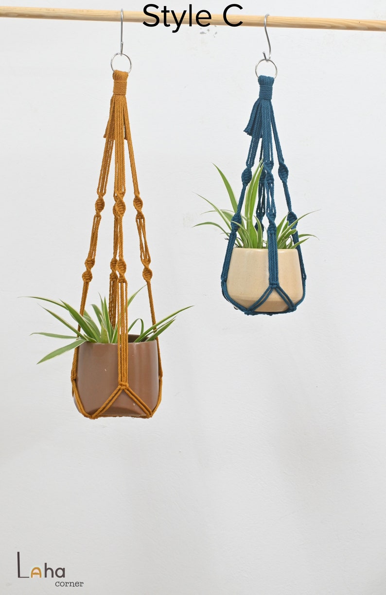 May include: Two macrame plant hangers, one in mustard yellow and one in teal blue, hanging from a wooden dowel rod. Each hanger has a small potted plant inside.