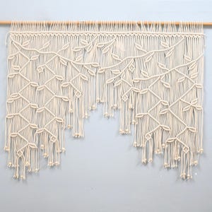 May include: An off-white macrame wall hanging suspended from a wooden dowel. The piece features an intricate knotted pattern resembling climbing vines with leaves, finished with long, loose fringe strands and small wooden beads at the ends.