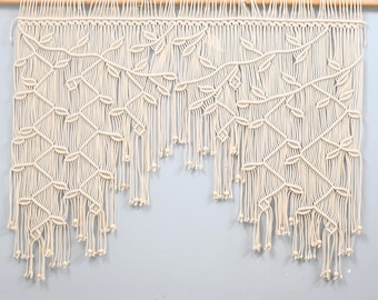 May include: An off-white macrame wall hanging suspended from a wooden dowel. The piece features an intricate knotted pattern resembling climbing vines with leaves, finished with long, loose fringe strands and small wooden beads at the ends.
