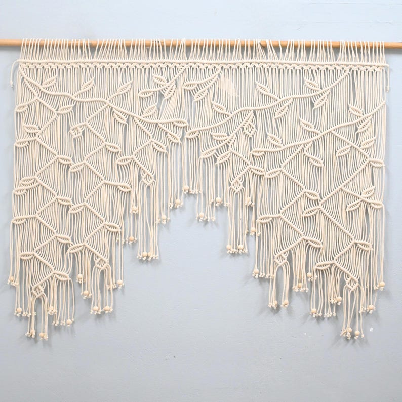 May include: An off-white macrame wall hanging suspended from a wooden dowel. The piece features an intricate knotted pattern resembling climbing vines with leaves, finished with long, loose fringe strands and small wooden beads at the ends.
