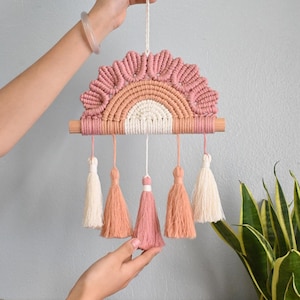 Puede incluir: Un colgante de pared de arcoíris de macramé hecho a mano en tonos rosa, melocotón y crema. El diseño presenta una barra de madera y borlas colgantes. Una pieza decorativa para guarderías o decoración de estilo bohemio.